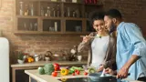 A husband and wife tasting food from a spoon in their kitchen. The counter is covered with colourful vegetables, such as peppers. loading=